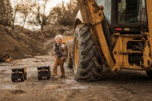 Two young boys dressed in play clothes excitedly push toy dump trucks over dirt mounds, fully immersed in their adventure.